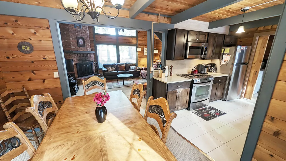 A wide view showing the open-concept living space, with a central fireplace, seating area, dining table for six, and the adjoining kitchen. Expansive windows allow natural light to flood the space, while vaulted wood ceilings add a spacious, airy feel. The warm color palette and cozy furnishings create a welcoming atmosphere ideal for gathering and unwinding.