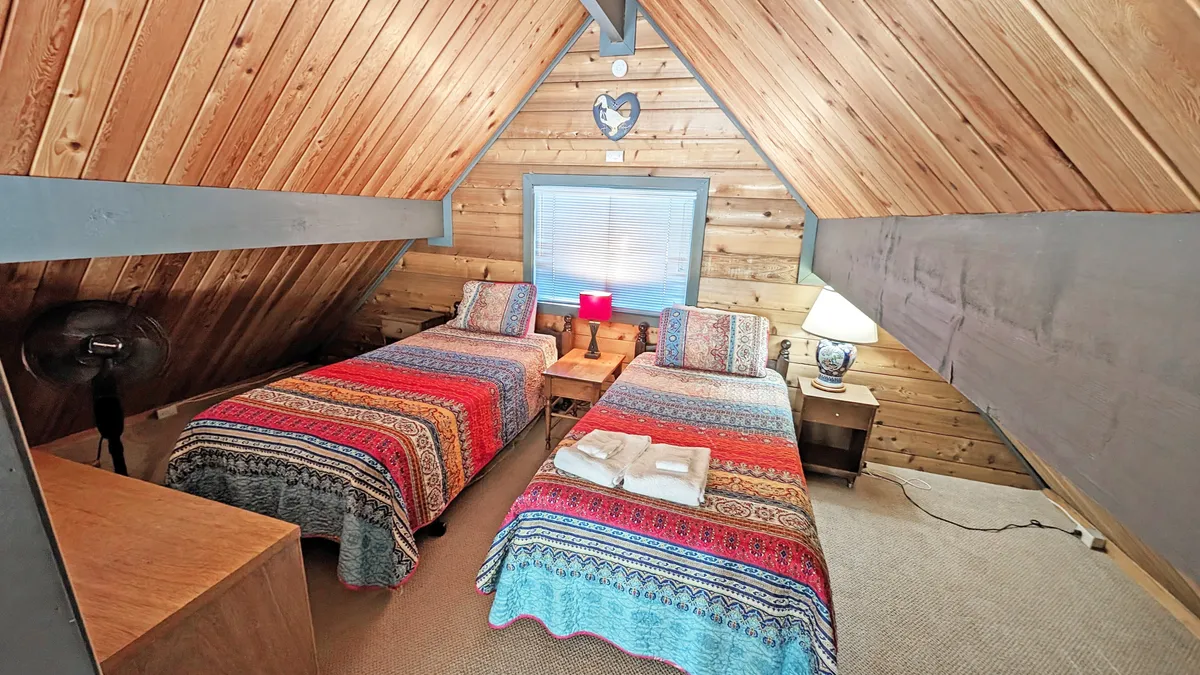 The airy loft features two twin beds with playful, colorful blankets beneath a wooden ceiling. A bedside lamp offers a gentle glow, making it a welcoming space for kids, teens, or friends seeking a fun and social sleeping nook.