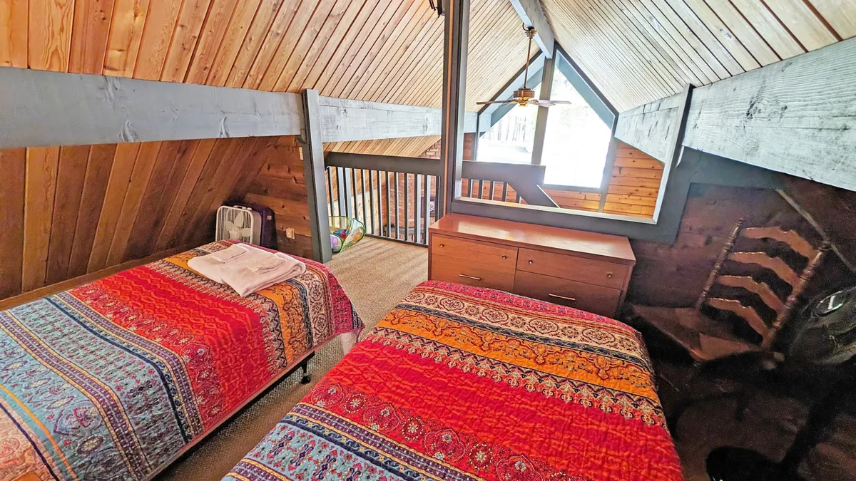 This photo showcases a loft or bedroom area with colorful bedspreads, a distinctive wooden ceiling, and large windows offering abundant natural light. The setting evokes a sense of warmth and togetherness, ideal for retreats with friends or a partner.