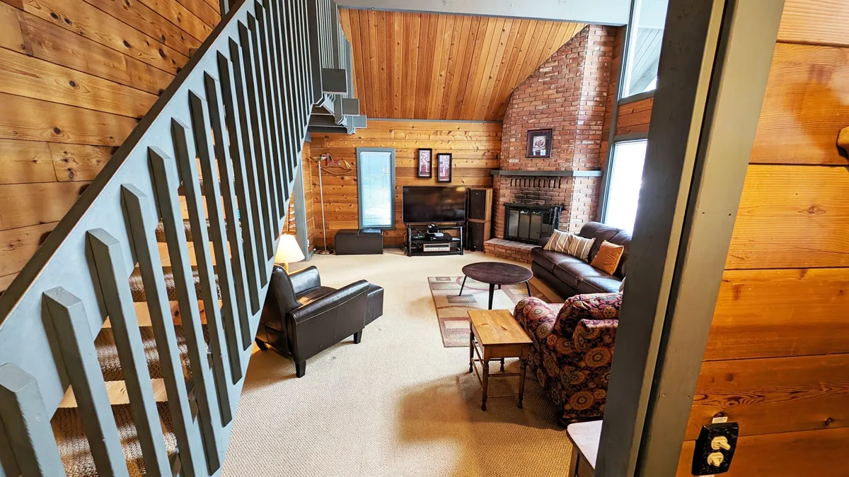 A warm and inviting living area with a fireplace, a comfortable sofa set, and wood paneling. The space feels open and connected, ideal for gathering after a day outdoors. The large windows (not visible but implied by the natural light and setting) likely offer glimpses of the forested surroundings.