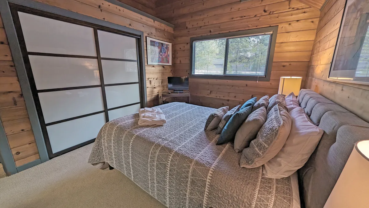 This warmly lit bedroom features a plush queen bed with soft pillows and quality linens, a large sliding glass door that lets in natural light, wooden wall paneling, and tasteful decorative artwork. The cozy vibe makes it perfect for unwinding after a day of adventure.