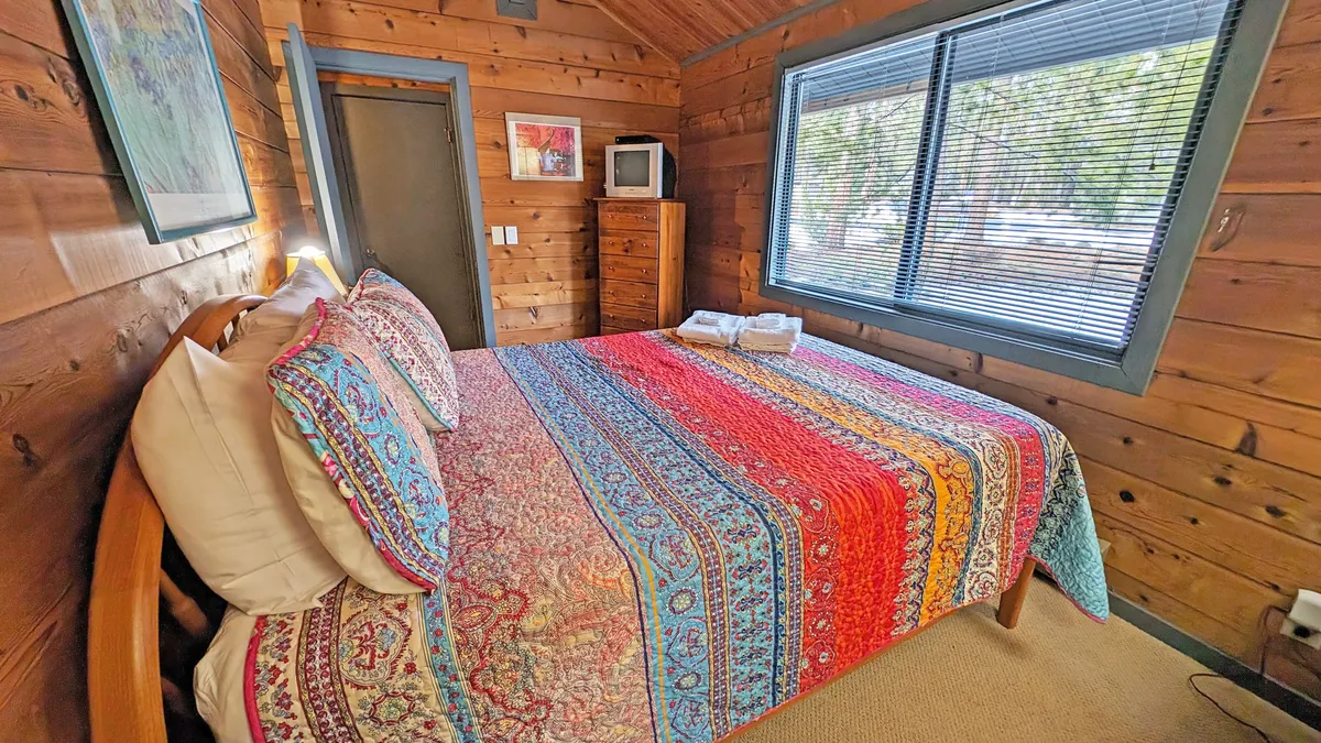 A charming bedroom with a queen bed dressed in colorful bedding sits against large windows framed by warm wooden walls, complemented by a dresser and a TV. Natural sunlight streams in, enhancing the sense of warmth and relaxation in the space.