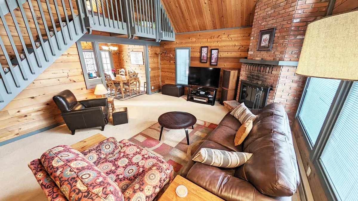 A cozy living space with a plush sofa, decorative pillows, a brick fireplace, and a TV unit. The dining area is visible in the background, illustrating the open layout. The scene radiates warmth and is set up for family relaxation, movie nights, or board games.