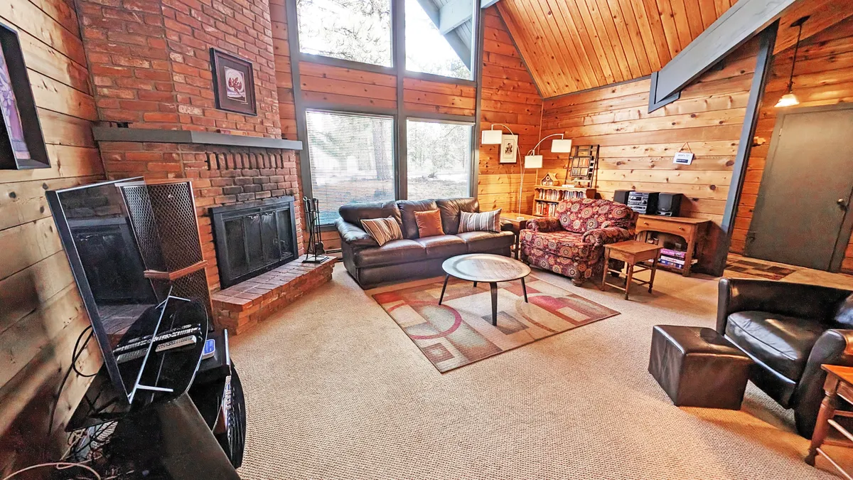 This image features a living space with large windows, a fireplace, and comfortable sofas arranged around a coffee table. The expansive windows allow natural light to flood in and provide views of the outdoors, suggesting a close connection to nature and the trails just beyond the home.
