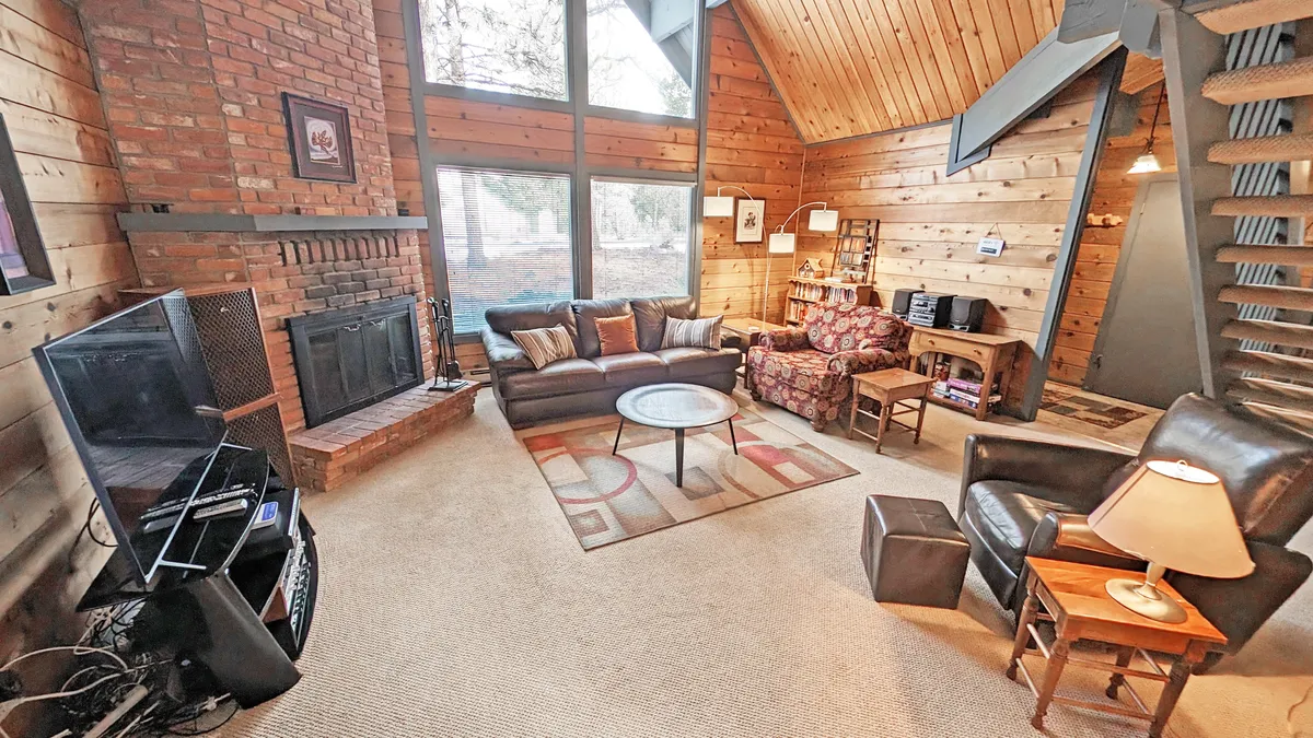The image showcases a bright, inviting living room with vaulted wood ceilings, large windows letting in natural light, a cozy fireplace, and plush seating arranged for relaxation or gathering. A bookshelf and games hint at leisurely evenings spent unwinding or playing together.