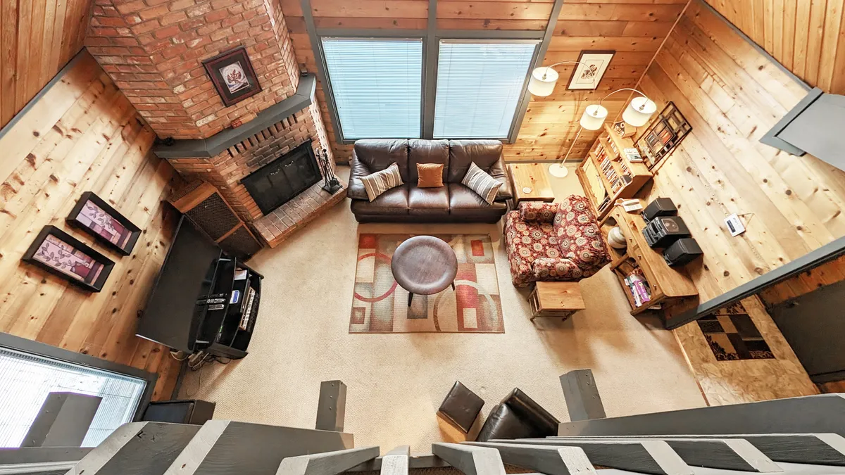 This image features a welcoming living area centered around a brick fireplace, complemented by a leather couch, a floral armchair, and wooden shelves. The large windows allow natural light to filter in and offer glimpses of the surrounding forest, creating a warm and inviting atmosphere.